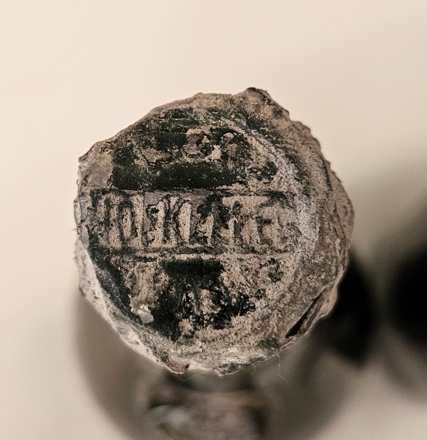 Metal capsule seen from above, struck with a lettered house mark across the centre