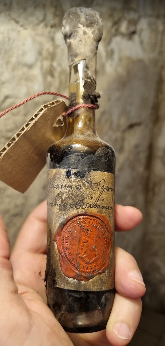 Small bottle with a handwritten label closed by a large red wax seal, a parchment tag corded to the neck
