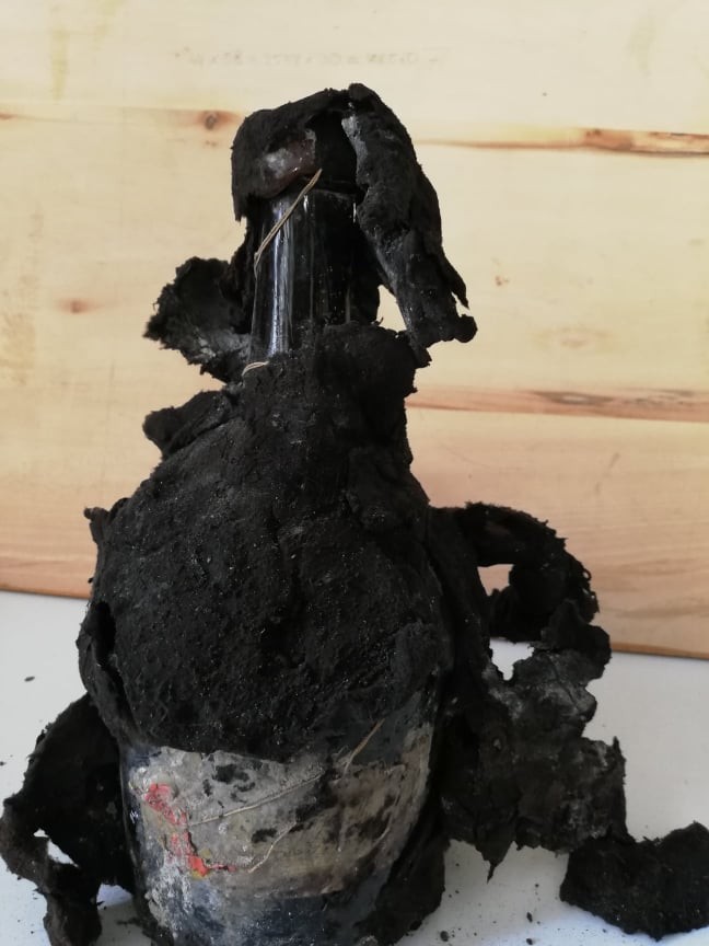 Bottle encased in a thick black crust of mould and wrapping, only the neck and a paper label showing