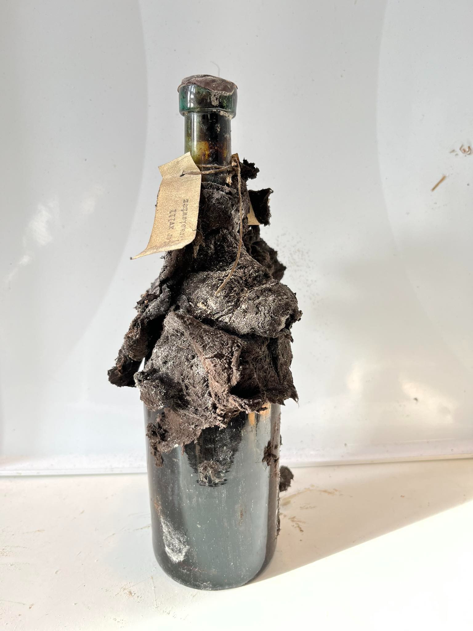 Bottle wrapped in blackened, mould-covered paper, neck strip still tied