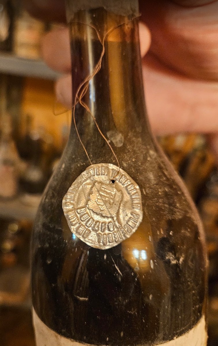 Embossed metal tag wired to the neck of a bottle, lettered BUDAFOK with a serial number