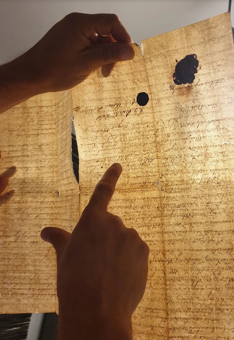 A document held up to the light, the writing legible through the sheet, with its black wax seal