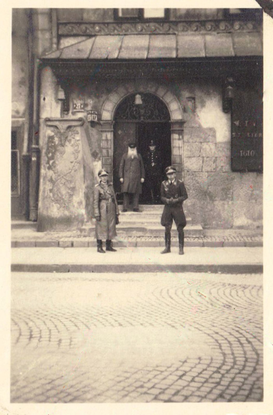 The Fukier shopfront under German occupation: two German officers on the pavement, an elderly bearded man and a Polish policeman in the doorway, the board reading T. Fukier 1610 at the right