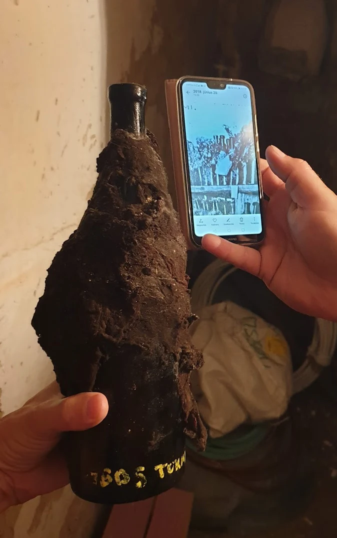 The encrusted neck of a bottle held up in the cellar where it was found, beside a phone showing a photograph of the walled-up chamber