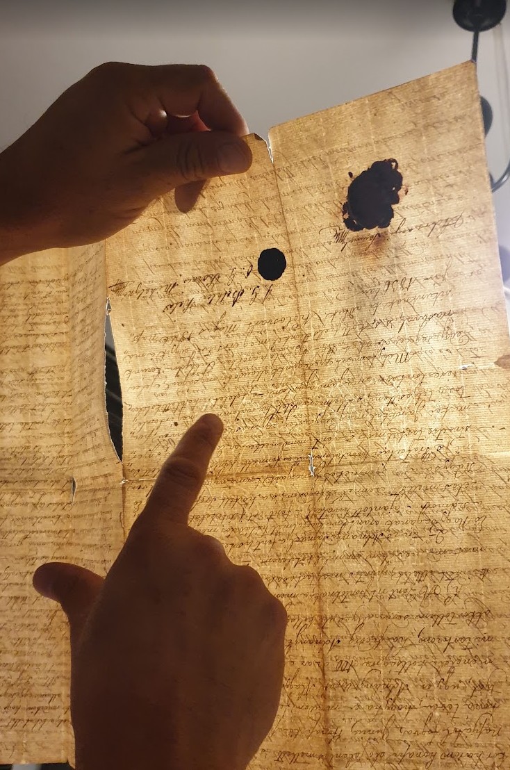 Handwritten document held against the light, showing the paper structure, a wax seal and a punched hole